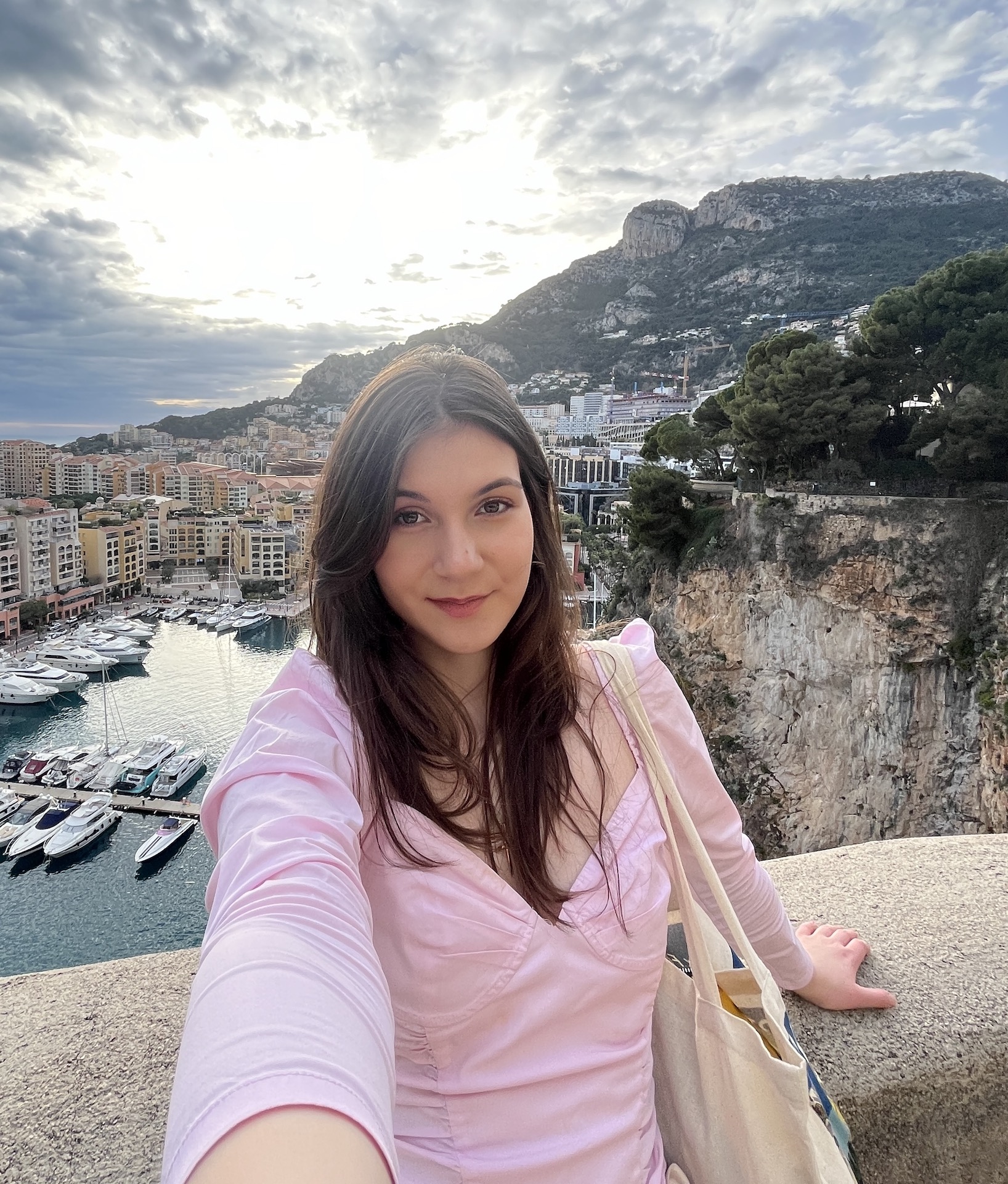 Tamar, a young woman with long brown hair, taking a selfie overlooking a harbour. In the background you can see boats, apartment blocks, and a hill overlooking the town.